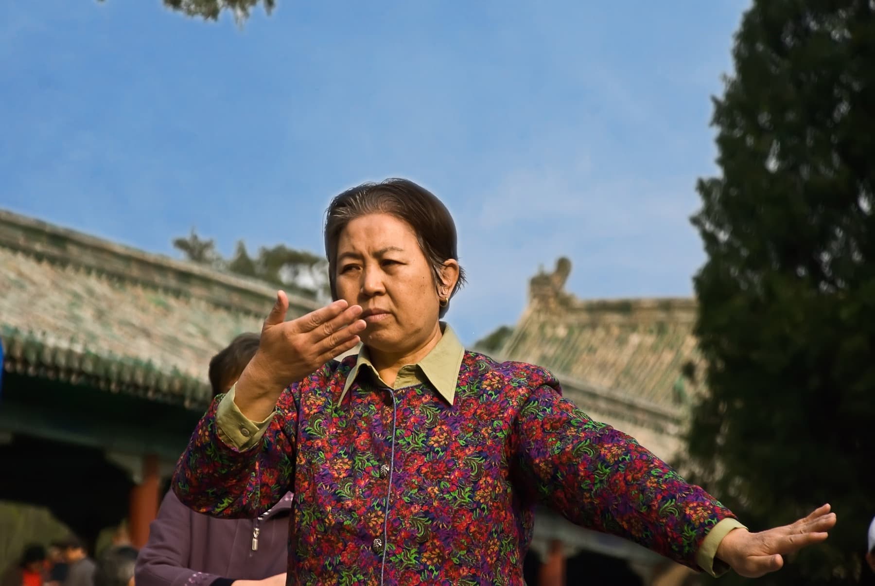 Older Chinese woman practicing tai chi outdoors, arms extended, in front of a traditional tiled roof