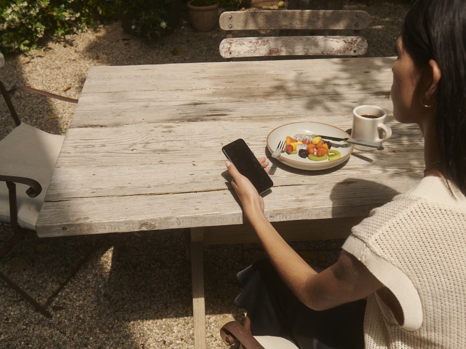 Young woman at an outdoor cafe table with coffee, fruit, and a phone in hand