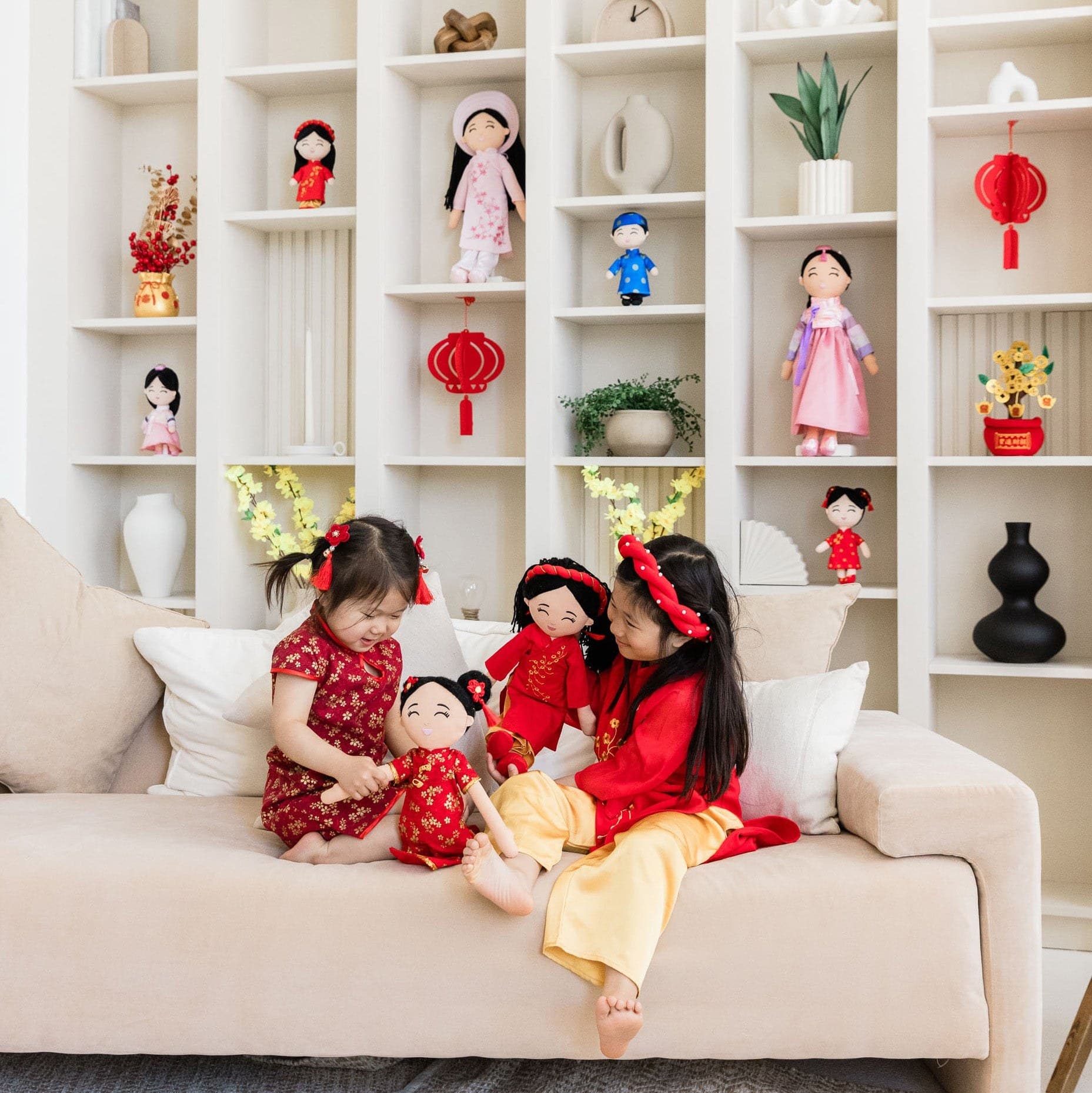Two Asian sisters in festive red outfits playing with culturally dressed plush dolls on a living-room couch